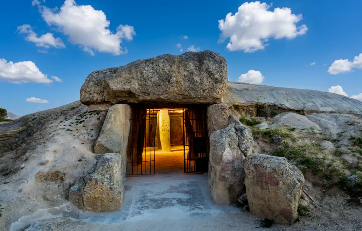 Open gates of the Dolmen of Menga revealing its lit interior and central stone pillar under a blue sky in Antequera