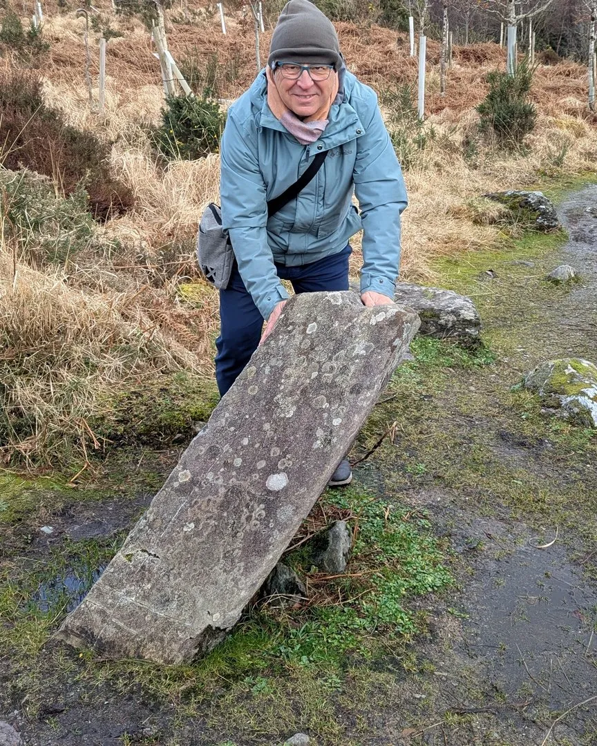 Carlos Rodriguez Silva con chaqueta azul y gorro gris sujetando un menhir de piedra en un sendero durante un tour innyou