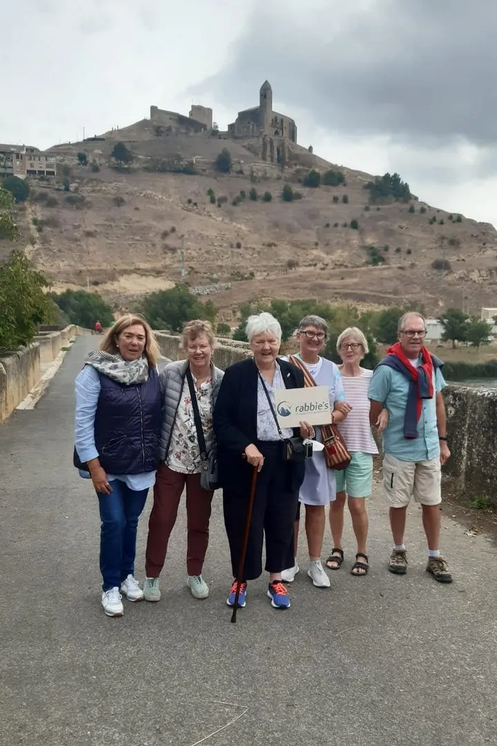 Grupo de turistas con Luz Bona Puerta posando en un puente con un castillo y una iglesia antigua sobre una colina al fondo. (SAN VICENTE DE SONSIERRA, GRUPO DE RABBIE’S TOURS 2023).